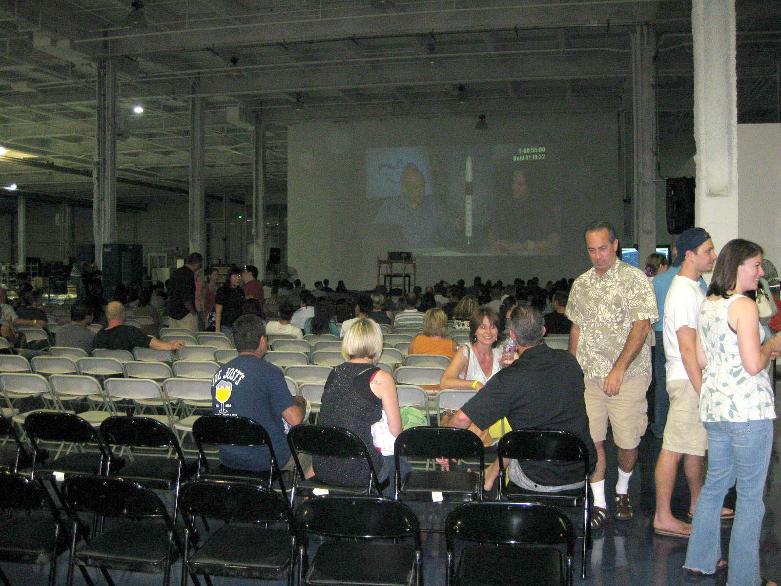 The Hawthorne hangar from the back: staff and families among rows of folding chairs, the webcast holding at T-55 minutes on the far wall.