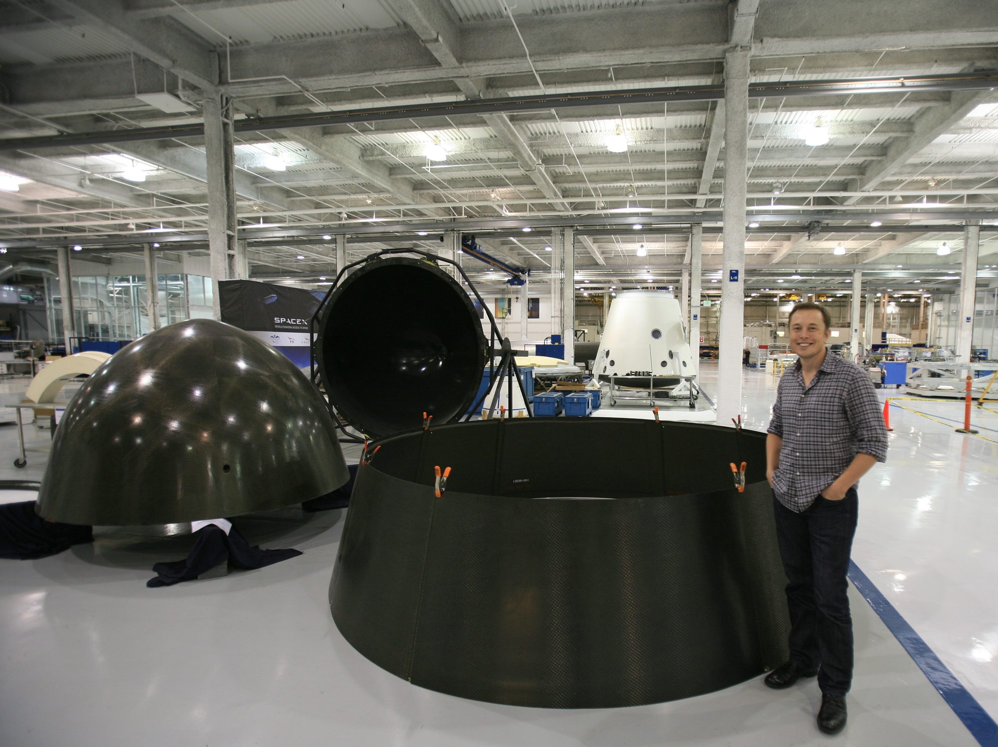 Elon Musk stands on the white factory floor at Hawthorne beside large black Falcon 9 tank domes and a ring section.