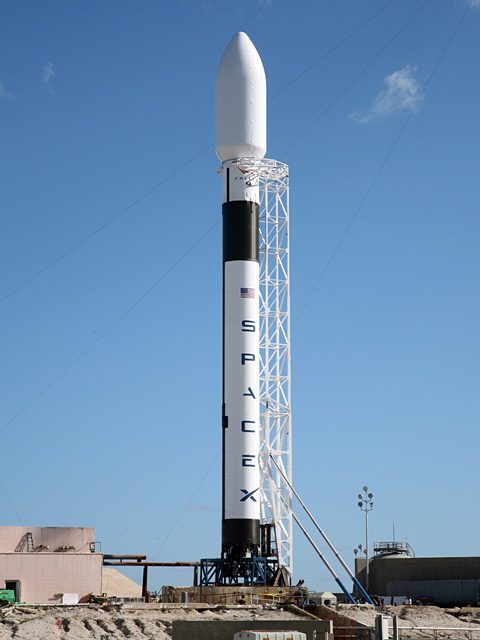 Falcon 9 standing in its white frame on the pad under a clear blue sky.