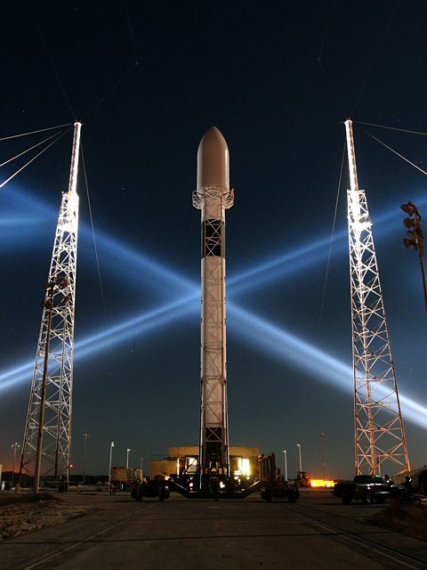 Falcon 9 at night on its pad between two lightning towers, two searchlight beams crossing in an X behind it.