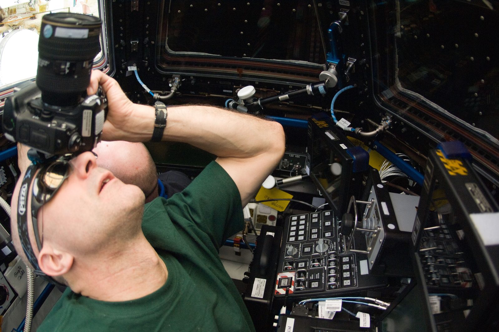 Don Pettit, in a green shirt, points a camera with a long lens up through a Cupola window.