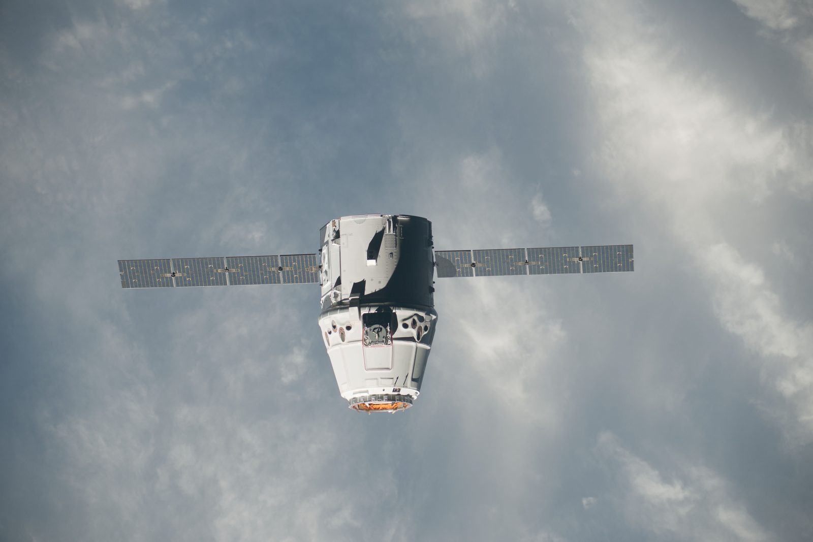 Dragon, nose toward the camera with both solar arrays spread, against white cloud.