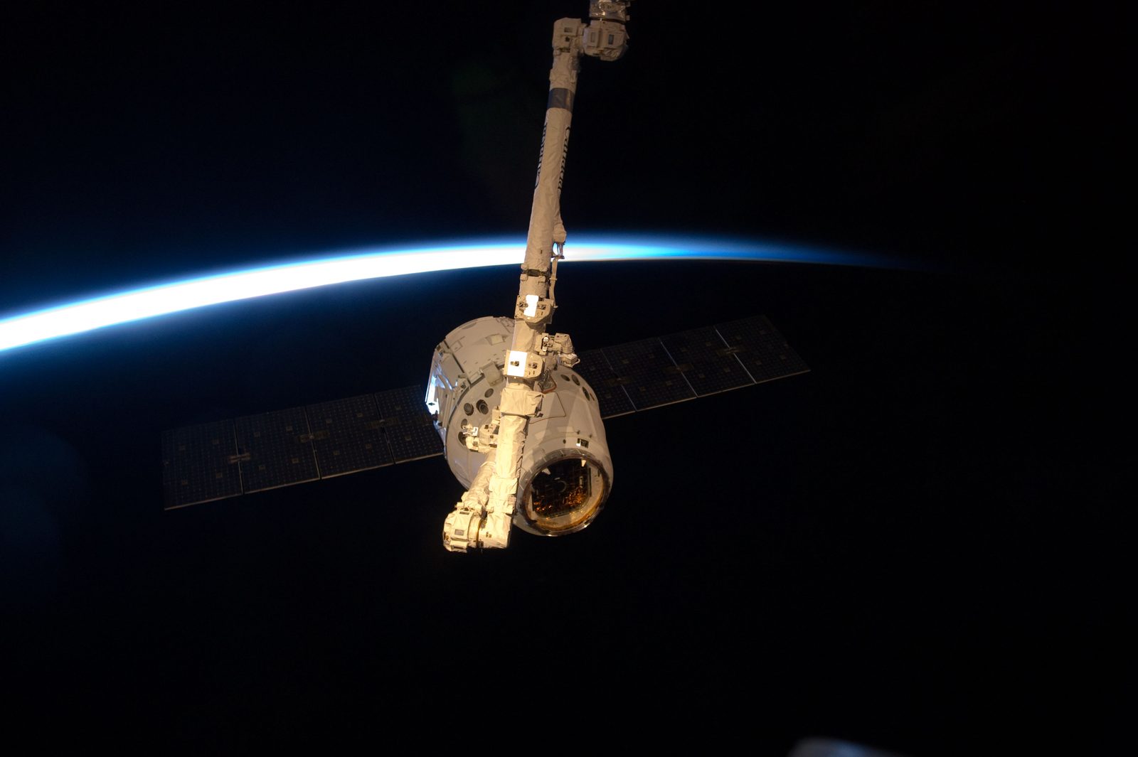 Canadarm2 holds Dragon against black space, with the thin blue line of Earth's atmosphere behind.