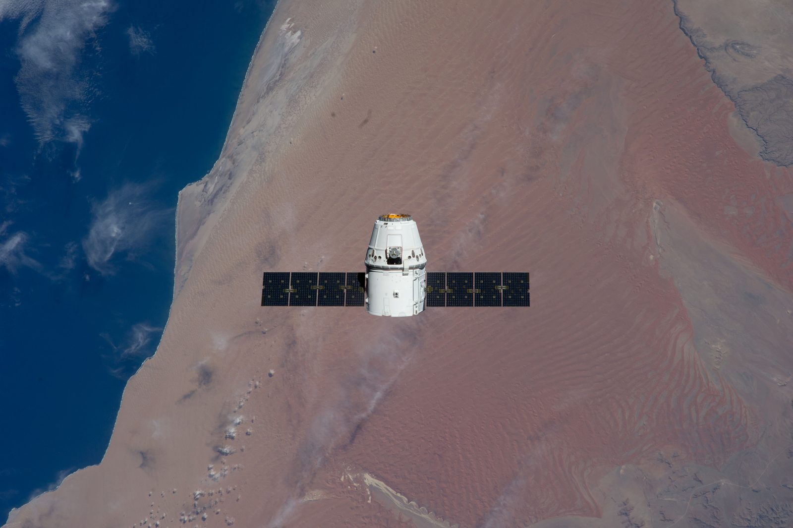 Dragon, small at center with its solar arrays spread, over the red sand of the Namib Desert and the Atlantic coast.