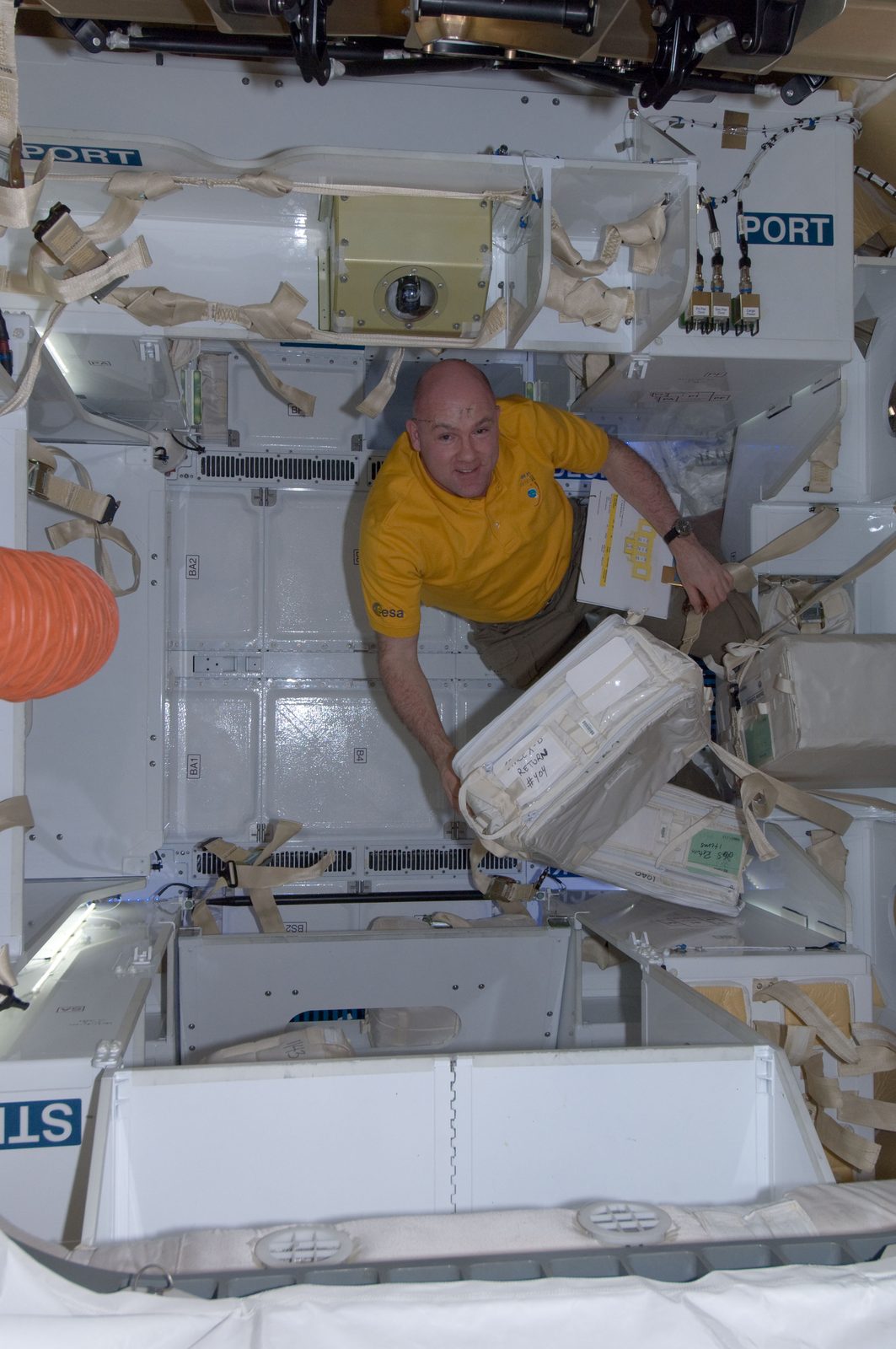 André Kuipers, in a yellow shirt, unpacks white cargo bags inside Dragon.