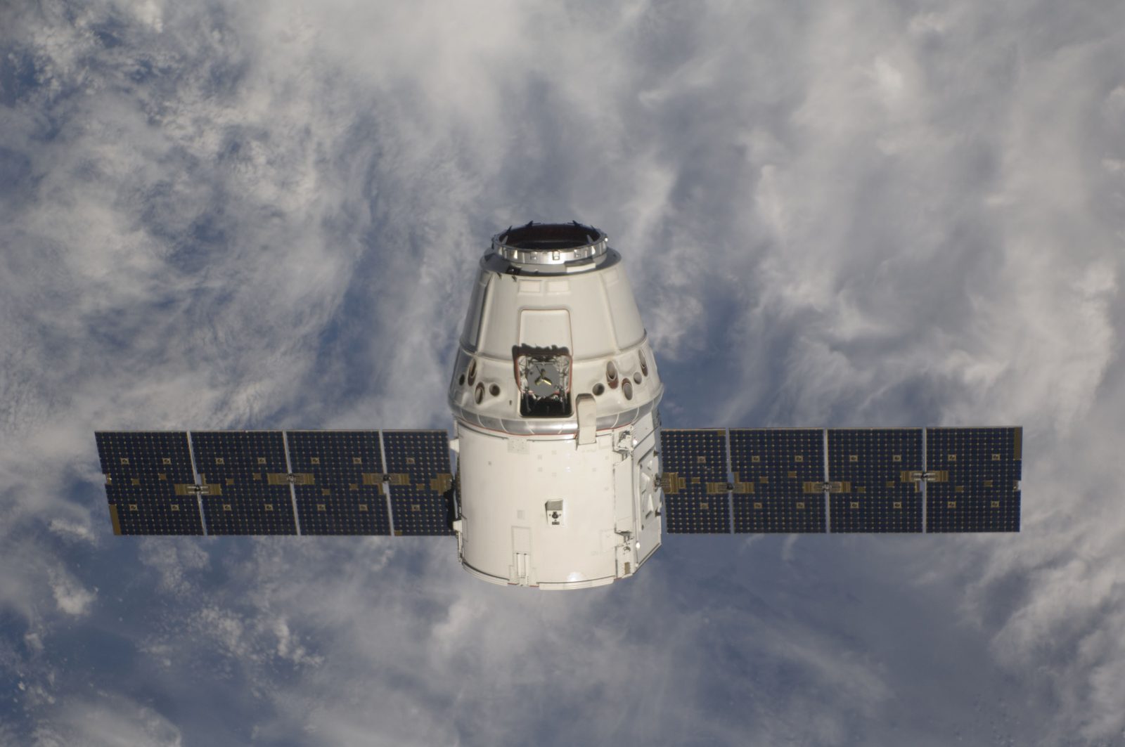 Dragon, nose toward the camera with both solar arrays spread, against cloud.
