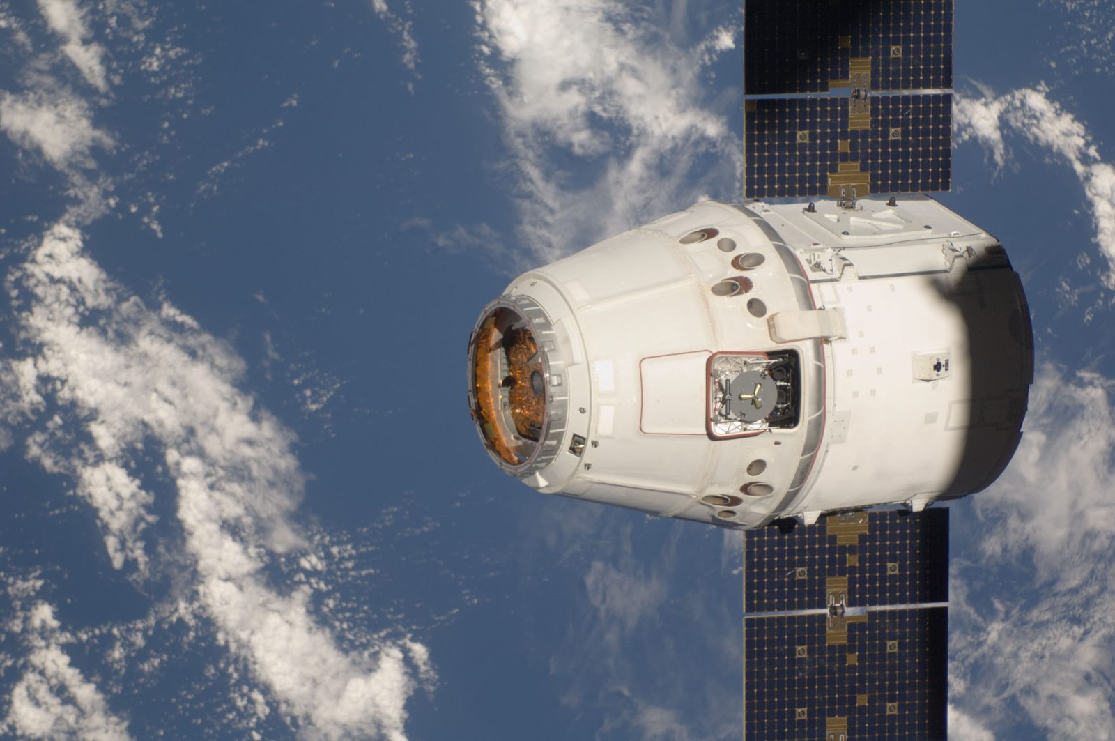Close side view of Dragon's capsule and grapple fixture, with a solar array behind and cloud and ocean below.