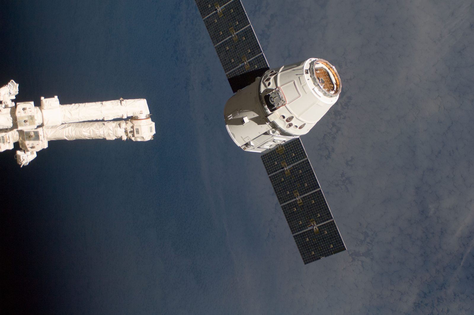 The end of Canadarm2 reaches in from the left toward Dragon, which floats over dark ocean.