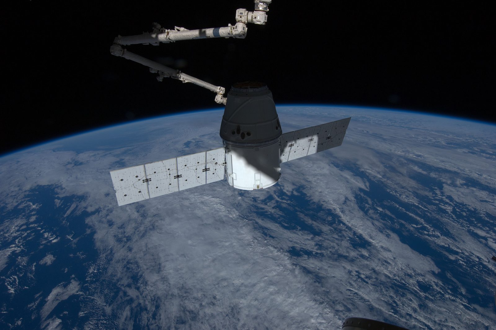 Canadarm2 holds Dragon over Earth, with the horizon and black space above.