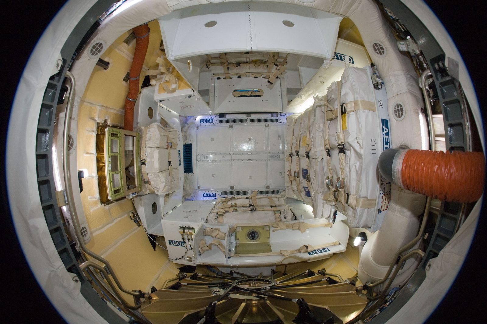 Fisheye view into Dragon's cabin: white cargo bags and racks, with an orange air duct at right.