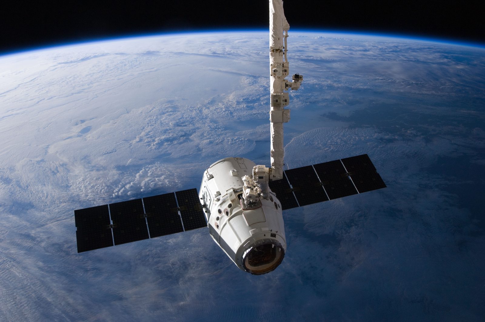 Canadarm2 holds Dragon, solar arrays spread, over Earth's curved horizon.
