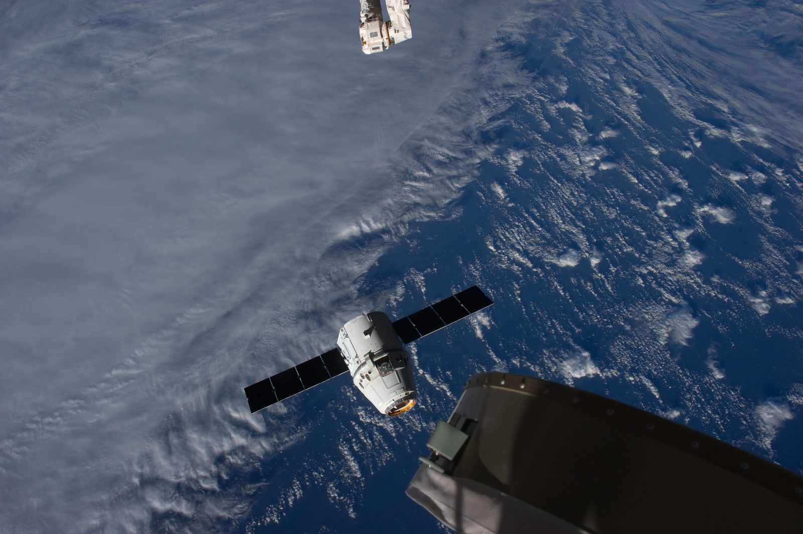 Dragon, free of the arm, drifts away over cloud-covered ocean; the arm's end is at top.