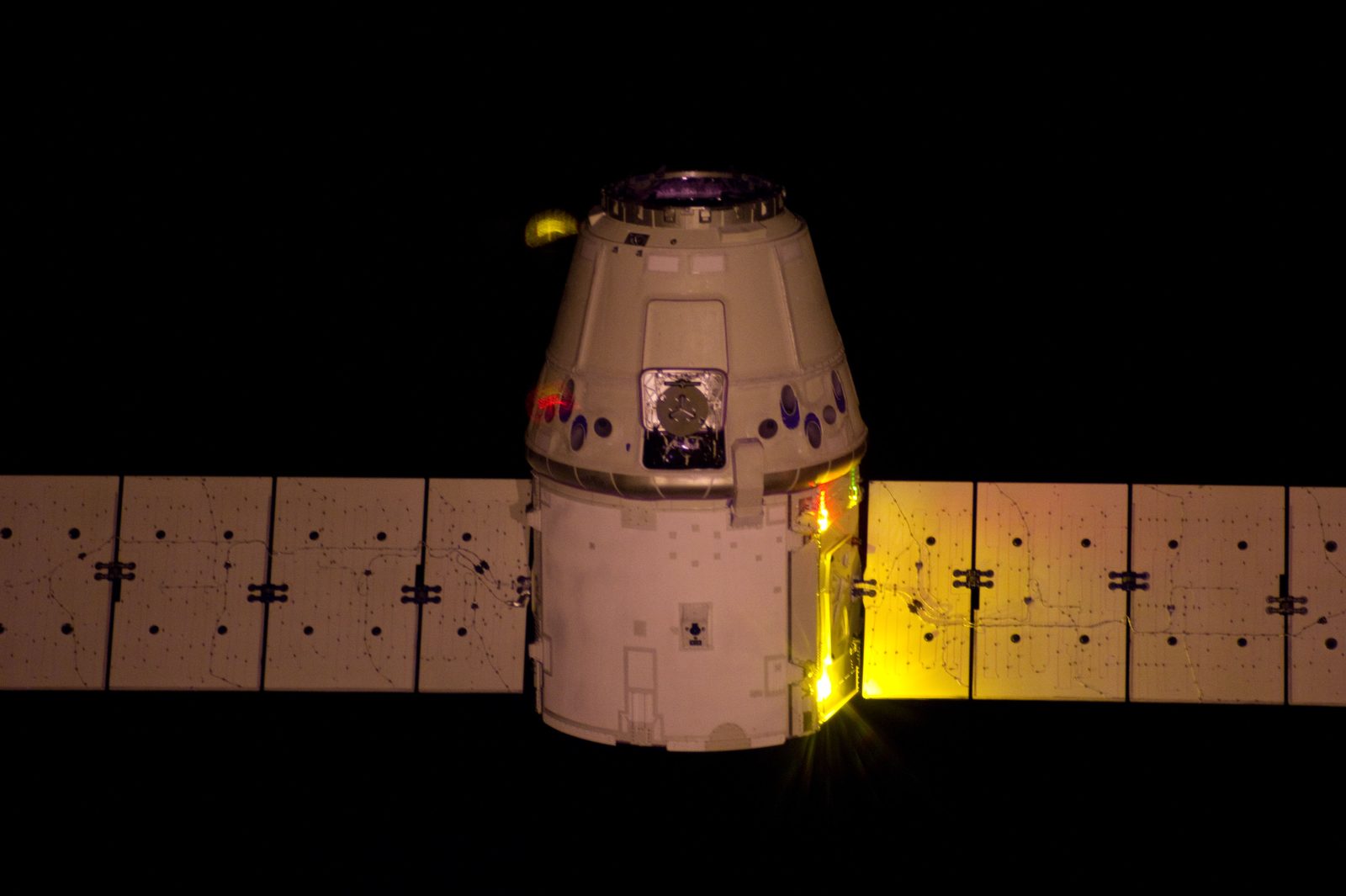 Dragon in darkness with its solar arrays spread, lit in warm tones by an infrared-modified camera.