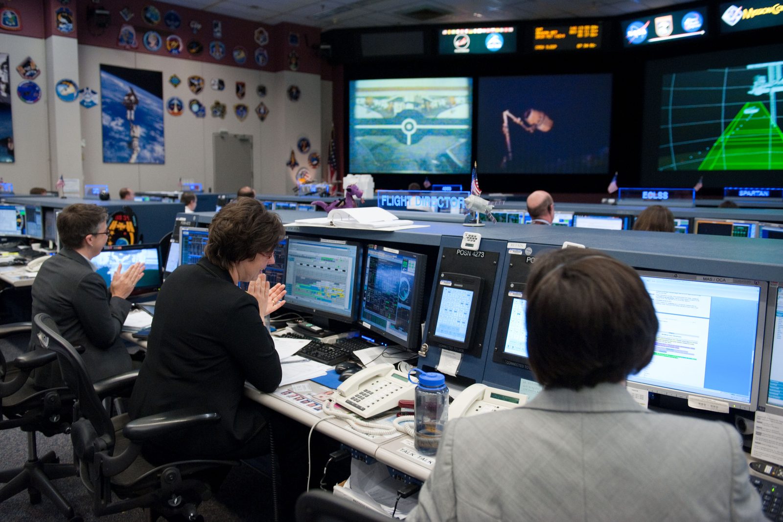 Flight controllers at a console in Houston applaud; the front screens show the arm holding Dragon.