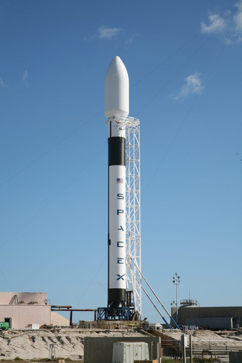 Falcon 9 stands vertical beside its launch tower at Space Launch Complex 40, under a clear blue sky.