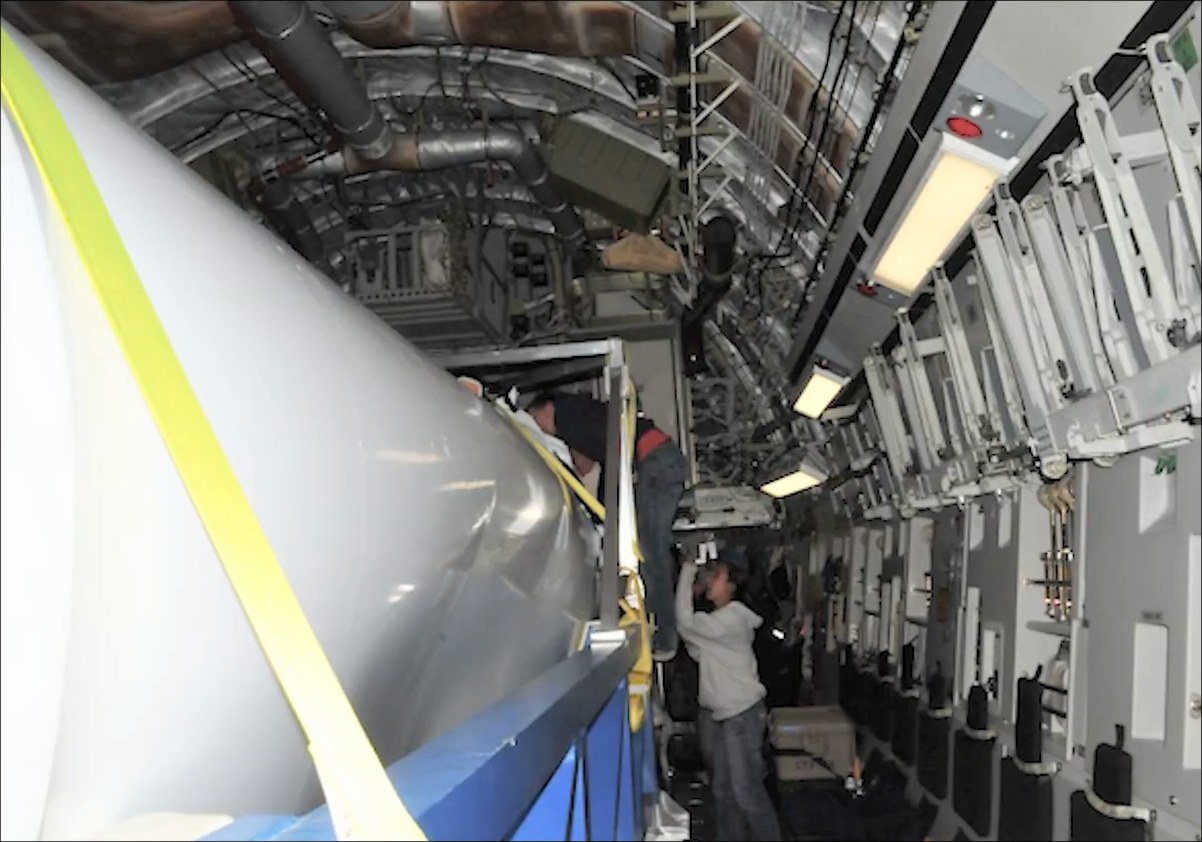 The white Falcon 1 first stage strapped down along the C-17’s hold, its tank wall sunk in and rippled; one person climbs in at the top of the stage while another stands below.
